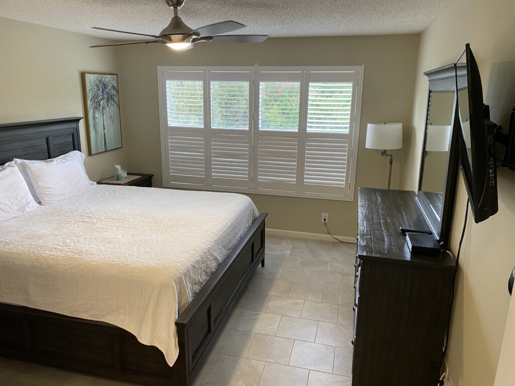 Primary bedroom displaying a wooden bedframe and dresser. Wood blinds cover the windows. There is a lamp in the corner, art on the walls and the floor is tiled.
